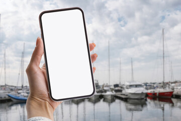 Female hand holding smartphone with white screen against marina background with yachts and sailboats under cloudy sky. Mockup for presentation and app design.