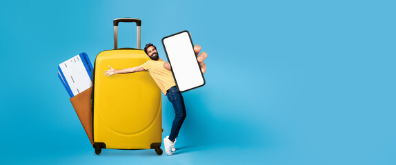 A man joyfully poses with a large yellow suitcase while holding a smartphone displaying a blank...