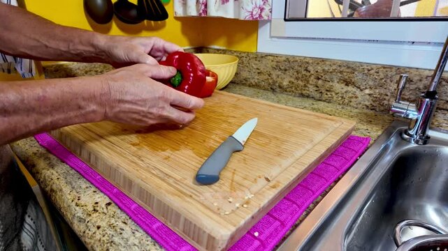 Experienced senior chef carefully removing seeds from ripe red bell pepper on wooden cutting board near kitchen sink, preparing fresh vegetarian ingredient