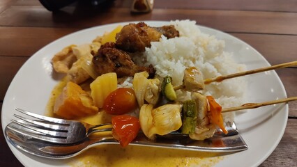 A white plate with a delicious-looking meal. With fluffy white rice and colorful vegetables and sliced meat . A fork and spoon are resting on the side of the plate.