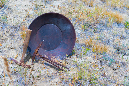 Gold-pan & mining picks rest in desert  showing landscape soil, mountain background.. Gold-pan is black metal. Digging picks are old & rusted. One is hand wrought metal or iron. Dig for gold invest?
