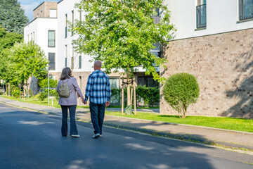 Happy Active mature couple walking hand in hand along urban street, suburban neighborhood, elderly man and woman enjoying walk together around residential, apartment buildings, modern architecture