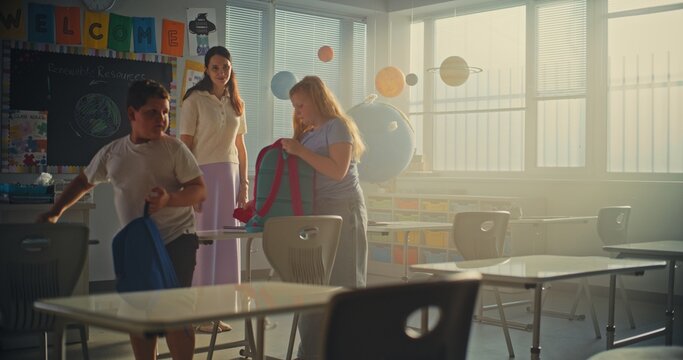 Female Teacher Finishing the Lesson, Announcing the End of School Day to Diverse Kids. Elementary School Students Standing Up, Packing Their Backpacks and Leaving Modern Classroom. Slow Motion. - Powered by Adobe