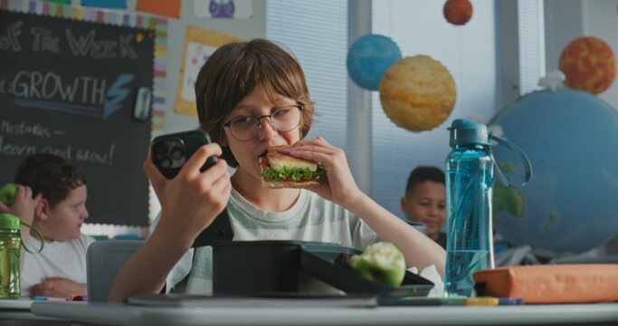 Elementary School Boy Sitting at the Desk, Eating Yummy Homemade Sandwich from Lunch Box, Using Smartphone. Young Boys and Girls Having Lunchtime in Classroom, Talking with Each Other Before Lesson.