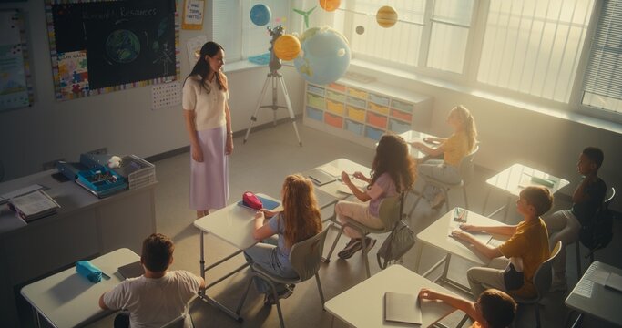 Female Teacher Finishing Lesson, Announcing the End of School Day to Diverse Kids. Elementary School Students Standing Up, Packing Their Backpacks and Leaving Modern Classroom. Slow Motion. High Angle