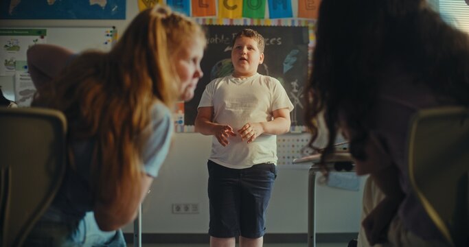 Primary School Boy Showcasing Knowledge of Ecology in Front of Class and Teacher. In Foreground, Two Girls Whispering During Environmental Science Lesson in Classroom. STEM Education. Slow Motion.