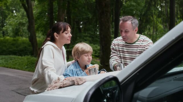 Young woman with red apple coming up to her husband and son during discussion of their route and location on paper map by car hook while standing on country road