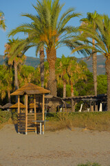 A thatched beach hut with a small table sits on a sandy shore, backed by blurred palm trees and distant hills under a clear blue sky.