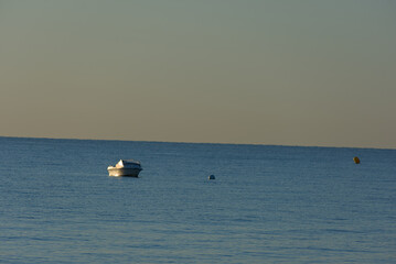 Fototapeta premium A small white motorboat with a light-colored canvas cover floats serenely on the calm blue water under a pale, muted sky.