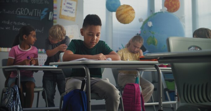 Stressed African American Primary School Boy Sitting Alone at Desk While Aggressive Classmates Abusing Him, Throwing Papers and Laughing. School Bullying, Racial Harassment, Toxic School Environment.