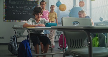 Depressed Primary School Boy Sitting Alone at Desk in Classroom While Aggressive Classmates Abusing...
