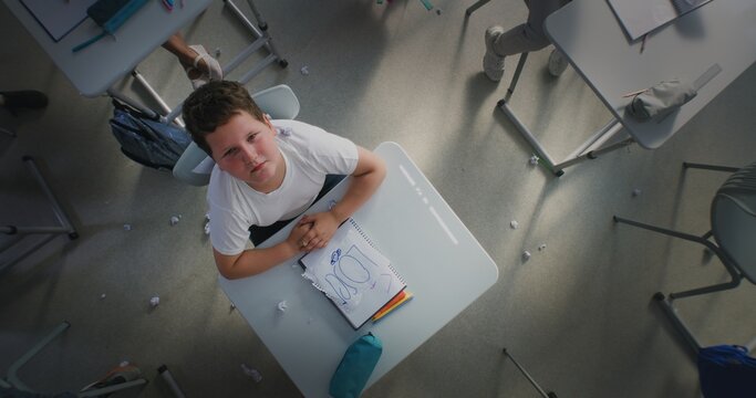 Depressed Primary School Boy Sitting at Desk, Looking at Camera While Aggressive Classmates Abusing Him, Throwing Papers and Laughing. School Bullying and Toxic School Environment. Rotating Top View. - Powered by Adobe