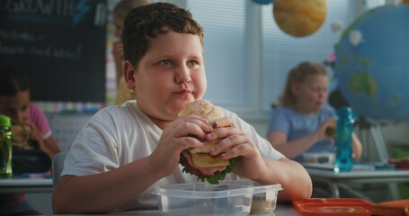 Elementary School Student Sitting at the Desk, Eating Yummy Homemade Sandwich from Lunch Box,...