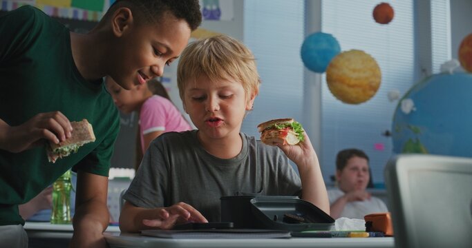 Primary School Boy Sitting at Desk, Eating Sandwich from Lunch Box, Using Smartphone and Talking to Classmate During Break. Happy Diverse Kids Having Lunchtime Before Lesson in Multiethnic Classroom. - Powered by Adobe
