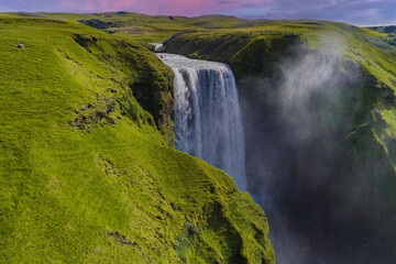 Skogafoss waterfall cascades into a misty gorge, surrounded by green cliffs and hills under a pink...
