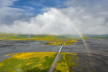 Aerial view of a straight road crossing a riverbed via a bridge in Iceland. A rainbow arcs over the...