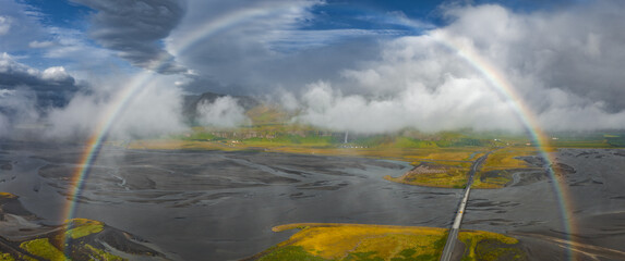 Aerial view of a braided river on volcanic sands in Iceland, with a double rainbow arching above. A road leads to hills under low hanging clouds.
