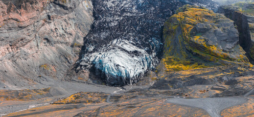 A glacier in Iceland surrounded by steep cliffs with volcanic rock, yellow moss, and a winding path leading to the icy blue and white glacier.