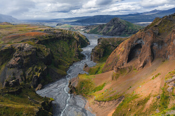 Mulagljufur Canyon in Iceland features steep cliffs, green vegetation, a winding river, rolling hills, and distant mountains under a partly cloudy sky.