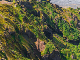 Lush green cliffs with steep rocky formations and deep crevices, set against a flat gray glacial...