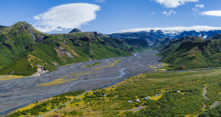 Obraz premium Aerial view of a green valley in Iceland with a braided river, rugged mountains, a glacier, and a campsite with vehicles and tents in the foreground.