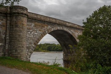 Historic stone bridge arching over a serene river under a cloudy sky in a natural landscape during autumn season