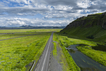 A paved road extends through a green landscape in Iceland, bordered by a stream and grassy...