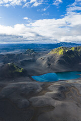 A vivid blue crater lake sits amid dark volcanic terrain in Iceland, with moss covered hills and mountains under soft sunlight and a partly cloudy sky. © Aerial Film Studio