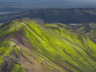 Vibrant green moss covered hills with dark volcanic rock, a dramatic ridge, and a flat plain in Iceland's unique volcanic terrain.