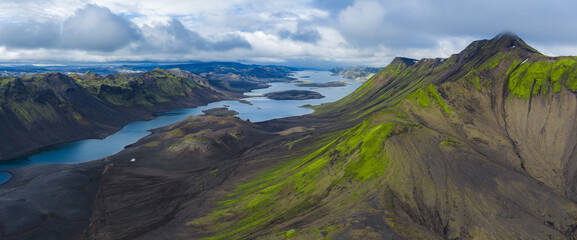 Aerial view of elongated lakes amid volcanic terrain and green moss covered mountains in Iceland, framed by dramatic cloud cover and sharp ridges.