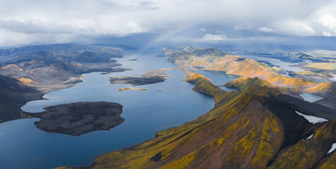 A vast lake in Iceland surrounded by rugged volcanic terrain with yellow and orange hills, deep blue water, and a faint rainbow under a cloudy sky.