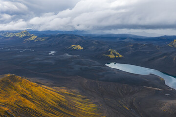 A dramatic aerial view of Iceland's volcanic terrain with dark rocks, yellow moss, a pale blue...