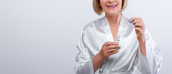 Cheerful senior lady in dressing gown using face serum on light studio background. Graceful mature...