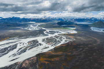 Braided rivers flow through a volcanic terrain in Iceland, with green patches, snow capped mountains, and rugged hills under a partly cloudy sky.