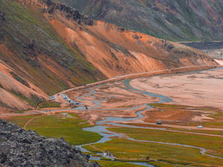 Vibrant orange and brown rhyolite hills with green mossy plains, winding streams, and a road with...