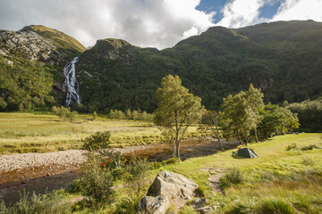 Scenic campsite by a waterfall amidst lush mountains and river in the highlands during daytime