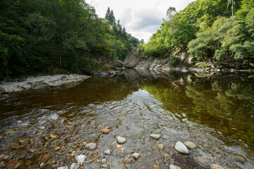 Calm river surrounded by lush greenery with rocky cliffs reflecting in the water under a cloudy sky