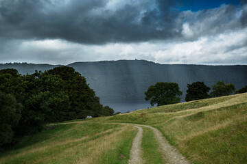 Scenic pathway through green hills leading to a body of water under a stormy sky near a forested mountain range