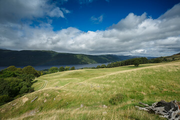 Expansive green landscape overlooking a tranquil lake surrounded by lush hills under a dynamic cloud-filled sky