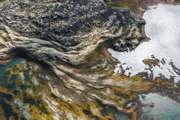 Aerial view of Iceland's volcanic terrain with dark rocky patterns, vibrant green moss, shallow water pools, and reflective surfaces on the right.