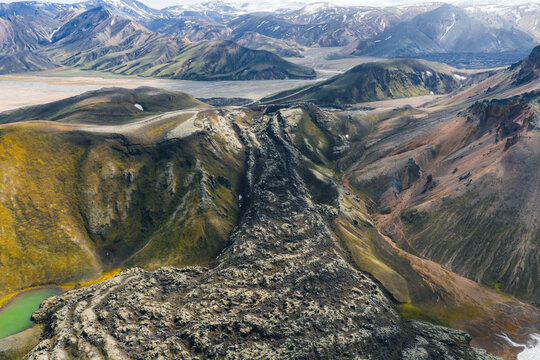 Aerial view of colorful rhyolite mountains in Landmannalaugar, Iceland, with a dark lava flow, patches of snow, and a small green lake in the foreground.