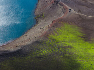 A vivid turquoise lake with a rugged reddish brown shoreline, bordered by green mossy terrain and...