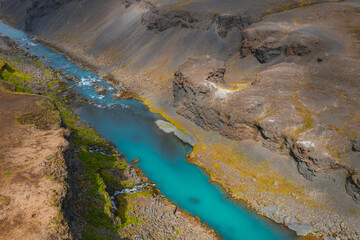 A vibrant turquoise river winds through a steep, rocky canyon in Iceland, with green moss and vegetation lining the rugged riverbanks.