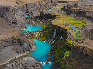 Turquoise waters flow through Sigoldugljufur Canyon in Iceland, with steep rock walls, cascading waterfalls, and green moss contrasting the rugged terrain.