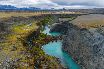 Sigoldugljufur Canyon features moss covered cliffs, cascading waterfalls, a turquoise river, rocky terrain, and distant snow capped mountains under cloudy skies.