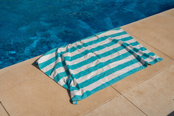 turquoise and white striped beach towel lying casually on the beige stone poolside edge, with...