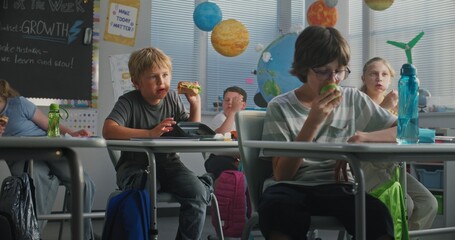 Primary School Children Sitting at the Desks, Eating Sandwiches from Lunch Boxes, Talking with Each...