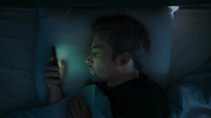 Close up, a tired man in bed scrolls endlessly on his mobile phone, struggling with insomnia, anxiety and overthinking as he watches social media late into the night. Smartphone dependence. Top view - Powered by Adobe