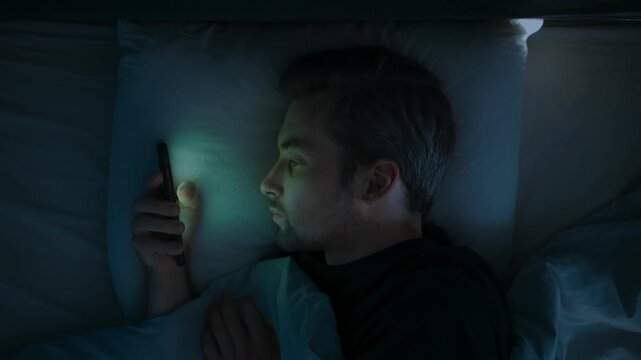 Close up, a tired man in bed scrolls endlessly on his mobile phone, struggling with insomnia, anxiety and overthinking as he watches social media late into the night. Smartphone dependence. Top view