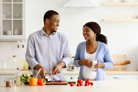 Positive expecting black woman and man cooking healthy food at home, looking at each other and smiling, kitchen interior. Happy african american pregnant couple making dinner together